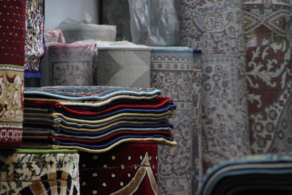 A close-up image of a neatly stacked pile of vintage and traditional patterned rugs, featuring intricate designs in shades of red, orange, beige, and blue. The rugs are folded and layered on top of ea