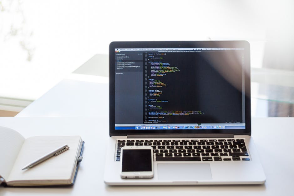 A close-up of a clean, modern desk featuring an open silver laptop displaying lines of programming code on a dark background, positioned centrally with the screen slightly angled towards the viewer. T